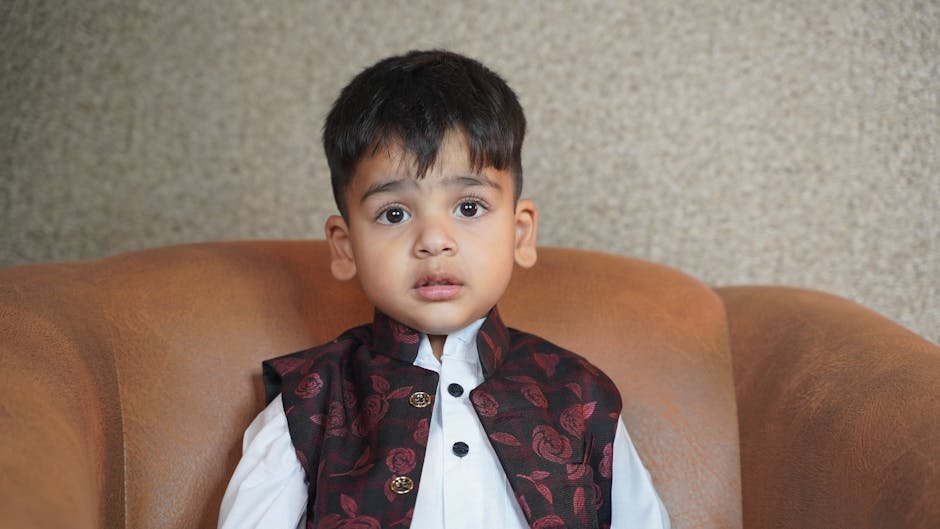 A young boy seated on a brown sofa wearing traditional attire, looking curious indoors.